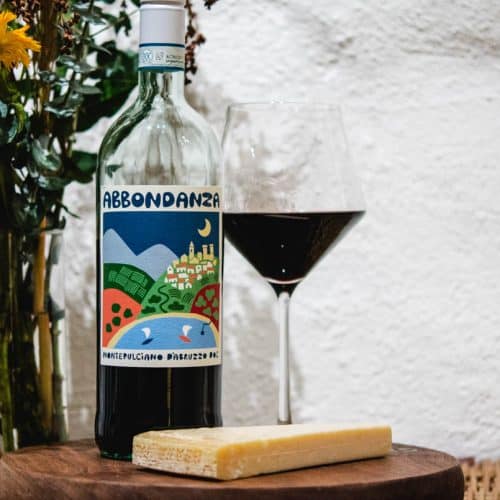 Abbondanza Montelpuciano D'Abruzzo wine bottle and glass with a wedge of cheese and flowers in the background