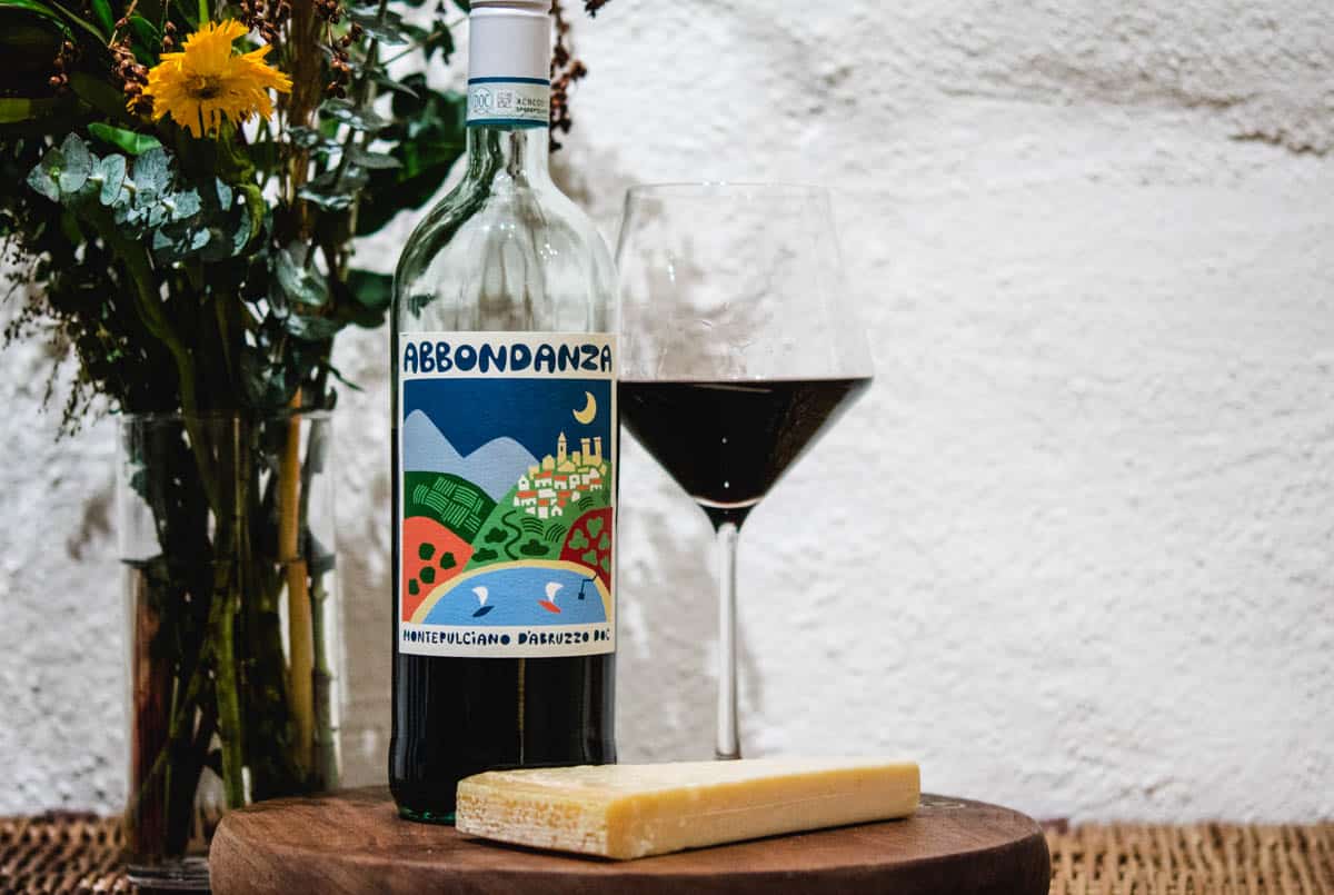Abbondanza Montelpuciano D'Abruzzo wine bottle and glass with a wedge of cheese and flowers in the background
