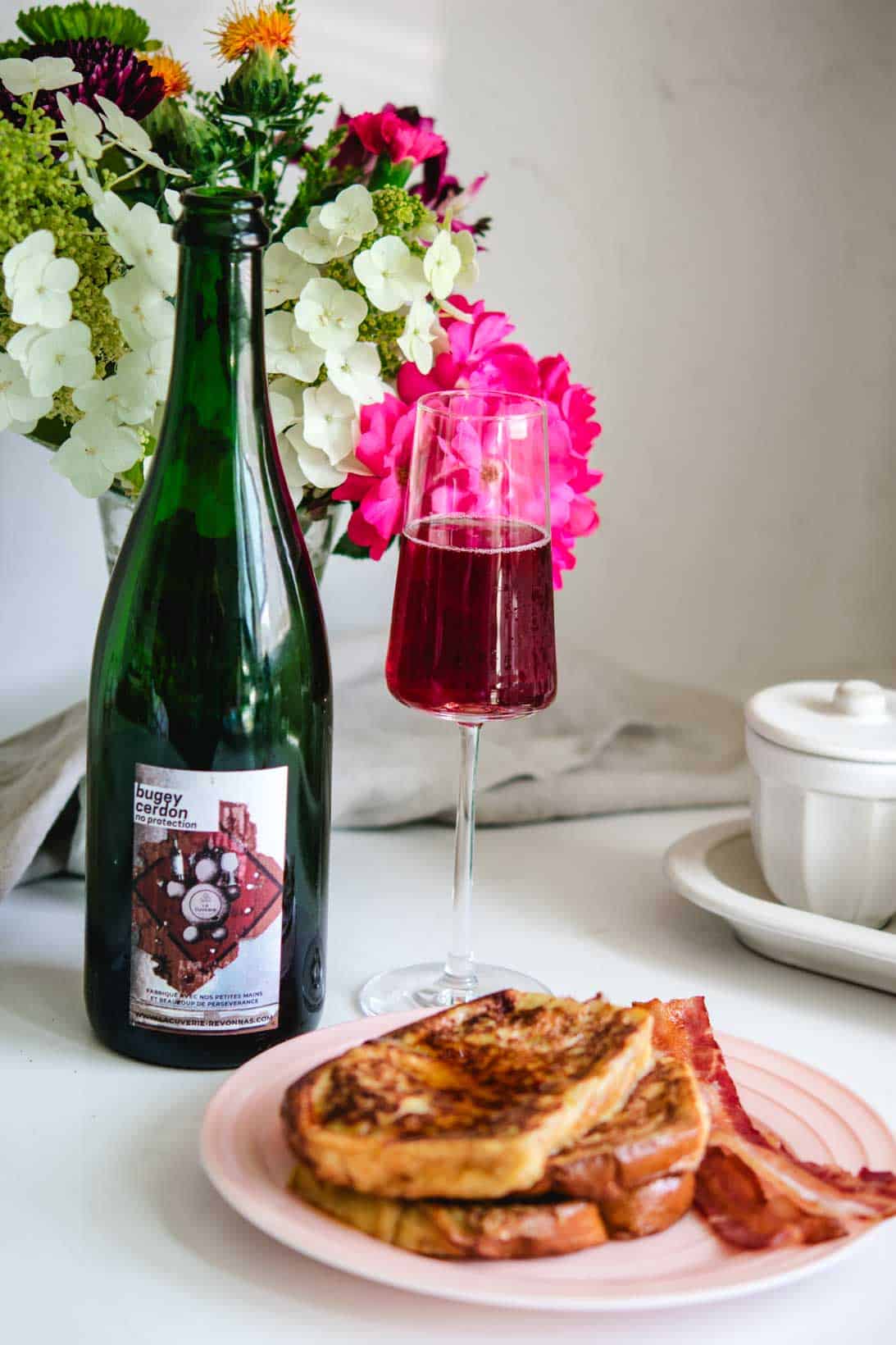 Wine bottle and glass of bugey cerdon with french toast and bacon with flowers in the background